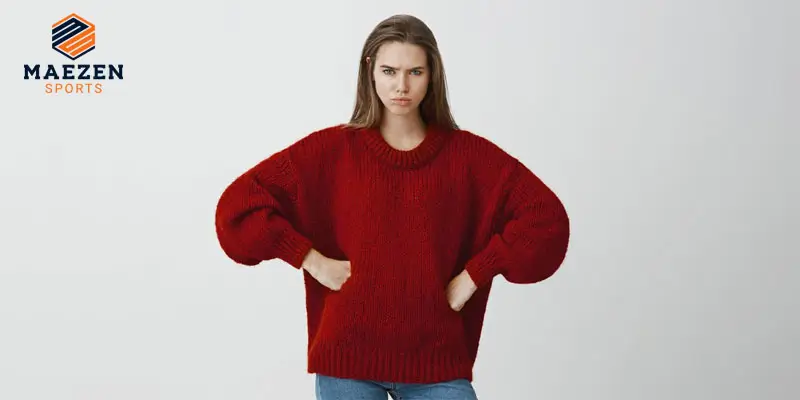 A woman wearing an oversized red sweater and jeans, standing with hands on hips, against a plain background.