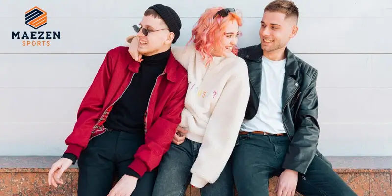  Three friends wearing streetwear, including a red jacket, white sweater, and leather jacket, sitting together and laughing against a white backdrop.