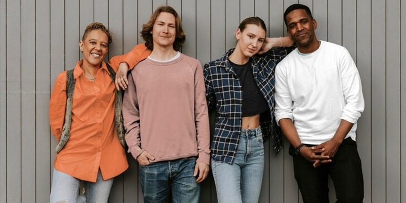 Four young adults standing together in casual outfits, smiling in front of a gray wall.
