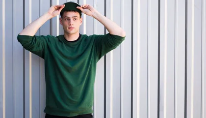 A man wearing a green sweatshirt and a baseball cap, posing against a modern industrial background, exuding casual streetwear style.