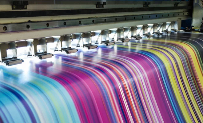 A close-up of a blue t-shirt featuring a coffee-themed design, prepared for printing with a screen printer while the artwork is being finalized on a computer.