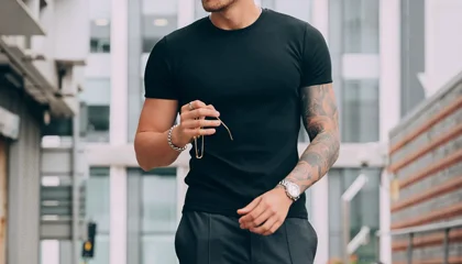 A man wearing a black t-shirt, paired with stylish trousers and accessories, walking confidently in an urban setting, showcasing a sleek and modern casual look.