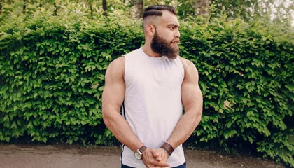 A muscular man wearing a white sleeveless t-shirt, showcasing his physique, standing outdoors in front of a green leafy background, perfect for fitness and active wear.