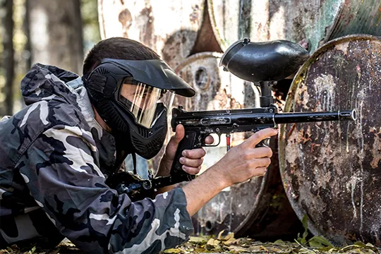 A paintball player in camouflage gear, kneeling behind barrels and aiming with a paintball gun, focused on the game.