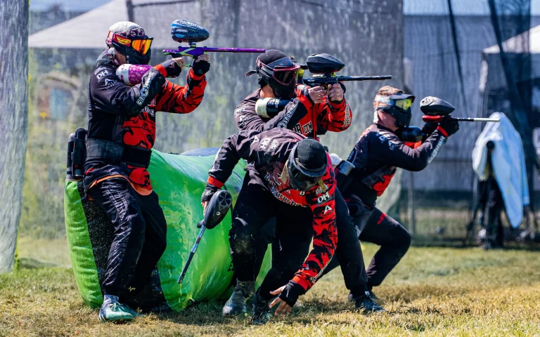 A group of paintball players in black and red jerseys, crouched behind cover, aiming and firing their paintball guns in a competitive match.