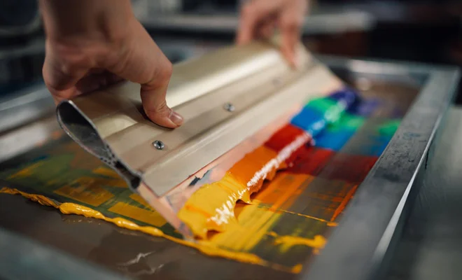 A close-up of a person using a squeegee to spread vibrant ink colors across a screen printing frame, demonstrating the screen printing technique for fabric design.