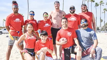 Group of people wearing red shirts posing for a photo on the beach during a flag football game.