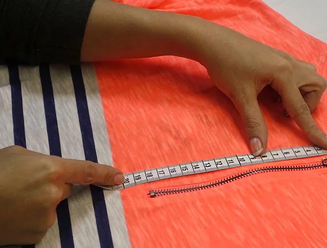 A close-up of a hand measuring the placement of a zipper on a garment using a tape measure. The shirt features vibrant orange and grey stripes, showcasing attention to detail in garment construction and fitting.