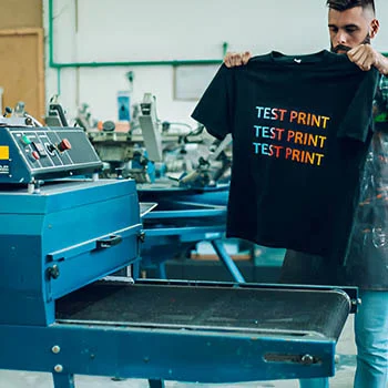 A person holding a t-shirt with 'TEST PRINT' in vibrant colors while working on a screen printing machine. shiw casing glow in dark print