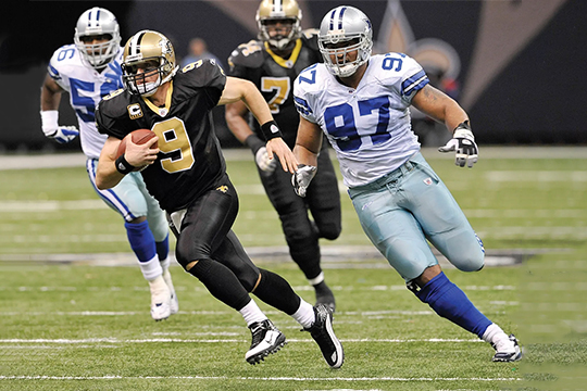 Action-packed moment from an American football game, with a quarterback in black and gold uniform evading a defender in blue and white, showcasing speed and skill on the field