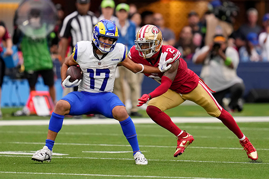 Intense American football action with a player in blue and yellow evading a defender in red and gold, showcasing the speed and athleticism of the game on the field.